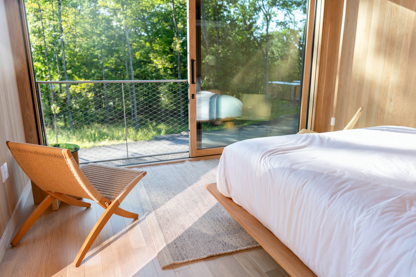 Moderna camera da letto con luce solare che filtra sul pavimento in legno e vista sulla foresta.