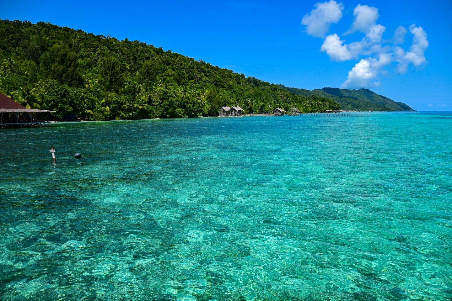 Bungalow di legno sopra l'acqua turchese limpida vicino a un'isola tropicale.
